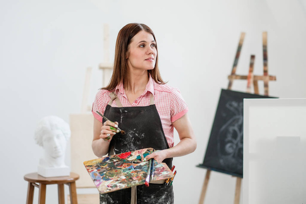 A young woman painter in a bright white studio draws a picture on canvas on an easel. Pink checked shirt, black apron, on the background of the pattern and tools. - Foto, Bild