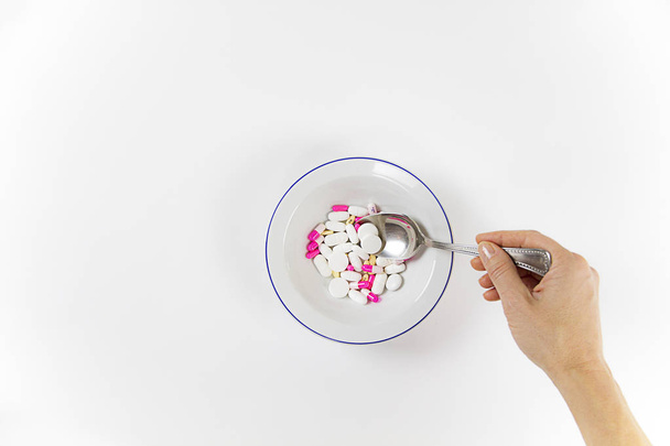 Пристрастие к лекарствам и наркотикам в наше время: рука молодой женщины с ложкой таблеток, взятых из тарелки, полной лекарств на белом столе и фоне
 - Фото, изображение