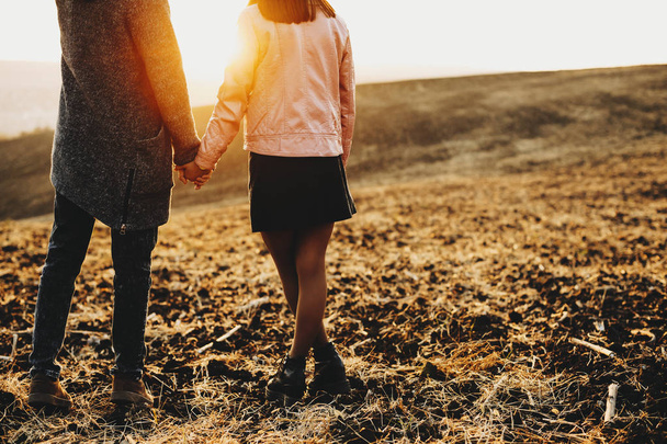 Zadní pohled na oříznutí muž a žena držící ruce přitom stát na poli trávě během nádherný západ slunce v krajině. Oříznout pár stojící v poli večer - Fotografie, Obrázek