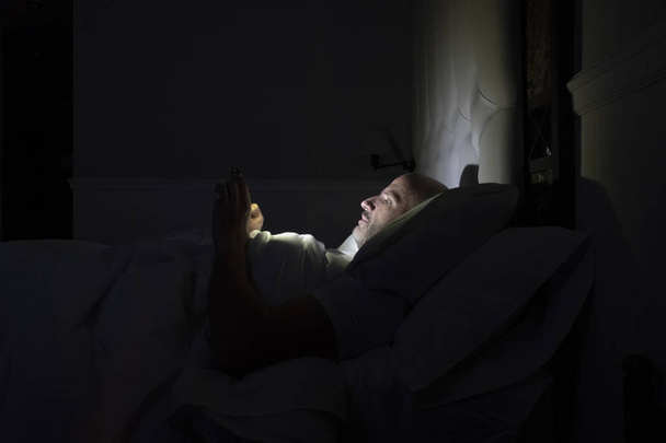 Ο άνθρωπος χρησιμοποιεί το τηλέφωνό του στο κρεβάτι του, αντί να κοιμάται, έννοια της τεχνολογικής εξάρτησης - Φωτογραφία, εικόνα