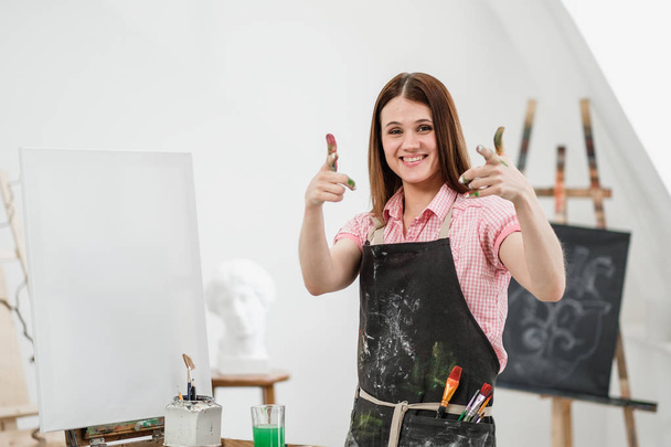 Mladá krásná dívka malíř v bílém studio s špinavé dlaně ukazuje třídu, se směje a jásá. - Fotografie, Obrázek