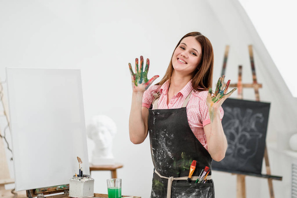 Young beautiful girl painter in a white studio with dirty palms shows class, laughs and rejoices. - Fotografie, Obrázek