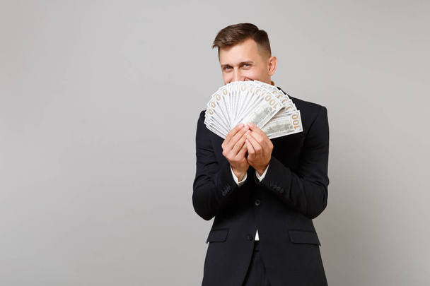 Cheerful young business man in suit hiding, covering face with lots bunch of dollars banknotes, cash money isolated on grey background. Achievement career wealth business concept. Mock up copy space - Fotó, kép