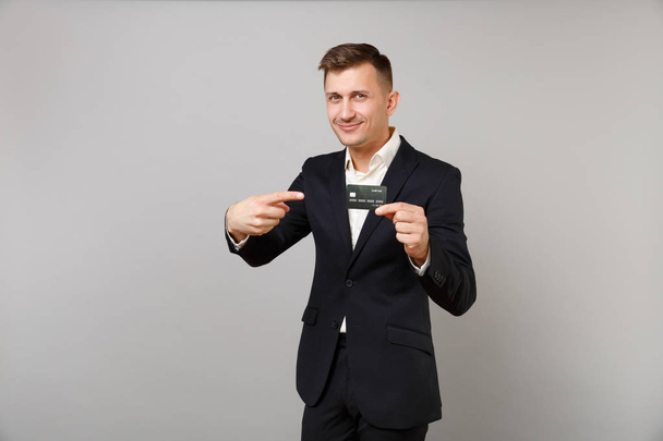 Smiling young business man in classic black suit pointing index finger on credit bank card in hand isolated on grey wall background. Achievement career wealth business concept. Mock up copy space - Photo, Image