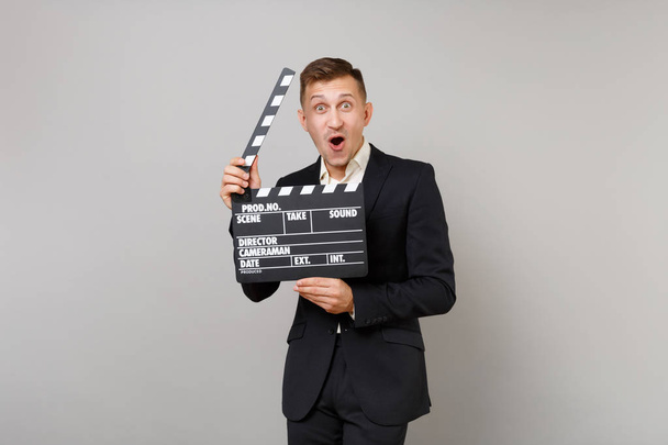 Excited business man keeping mouth wide open, looking surprised hold classic black film making clapperboard isolated on grey background. Achievement career wealth business concept. Mock up copy space - Photo, Image