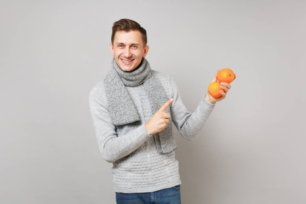 Jeune homme souriant en pull gris, écharpe pointant index sur oranges isolées sur fond mural gris. Mode de vie saine, émotions sincères, concept de saison froide. Maquette espace de copie
 - Photo, image