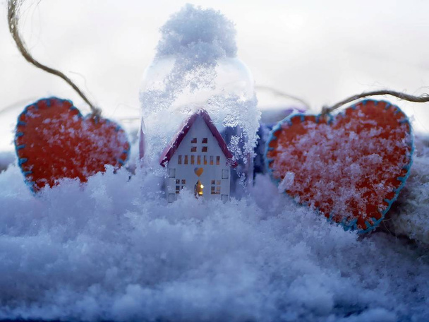 ein kleines weißes Spielzeughaus mit rotem Dach und beleuchteter Beleuchtung und einem Paar orangefarbener Filzherzen auf schneebedecktem Schnee, das Konzept der saisonalen Winterferien, Valentinstagsgrüße, Wohnkomfort - Foto, Bild