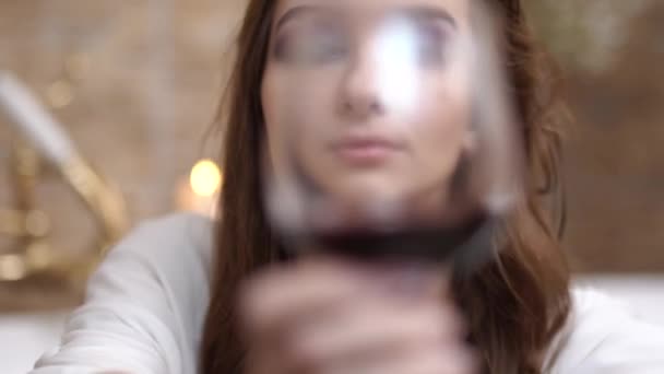 Portrait of a thoughtful girl taking a bath with bright makeup in a white shirt with wine glass. - Video