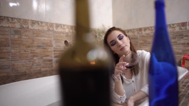 Portrait of a thoughtful woman taking a bath with bright makeup in a white shirt with wine glass. - Video