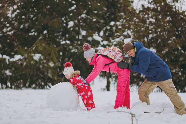 Family woman, man little girl in warm clothes playing, making snowman, pushing each other in snowy park or forest outdoors. Winter fun, leisure on holidays. Love relationship family lifestyle concept - Фото, изображение