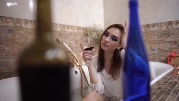 Portret van boos meisje nemen van een bad met lichte make-up in een wit overhemd met glas wijn. Flessen op de voorgrond aan de rand van het bad. - Video