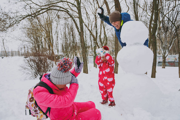 Cheery family man woman and little girl in warm clothes throwing snowball making snowman in park or forest outdoors. Winter fun, leisure on holidays. Love relationship family people lifestyle concept - Photo, Image