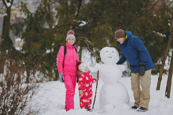 Cheerful young family man woman little girl in winter warm clothes standing near snowman in park or forest outdoors. Winter fun, leisure on holidays. Love relationship family people lifestyle concept - Photo, Image