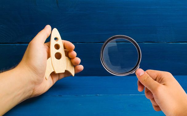 Magnifying glass is looking at the development of outer space. Blue wooden background. Training, game. Rocket in hand concept. The hand holds a wooden rocket. The rocket is in the way. Space camp. - Foto, Imagen