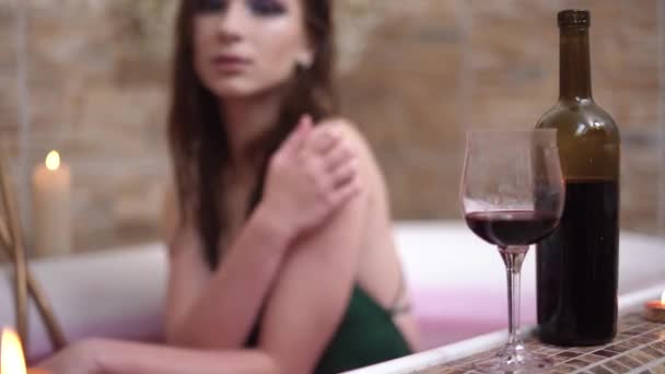 Bottle of red wine with glass are in bath edge. Smiling woman in the background sitting in bath - Footage, Video