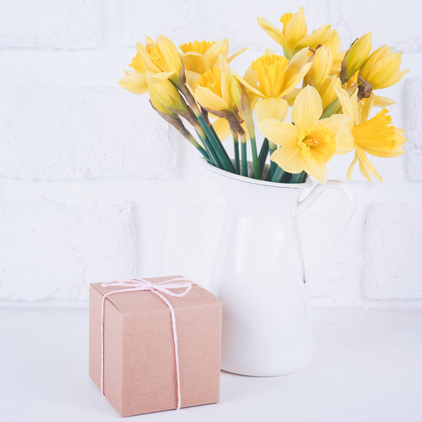 Frasco com narcisos e uma caixa de presente de papelão no fundo de tijolo branco. Primavera, dia das mães e conceito de Páscoa. Quadrado
 - Foto, Imagem