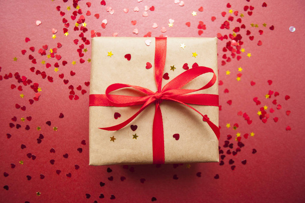Gift box wrapped in brown colored craft paper and tied with red bow on dark red background with red confetti. - 写真・画像