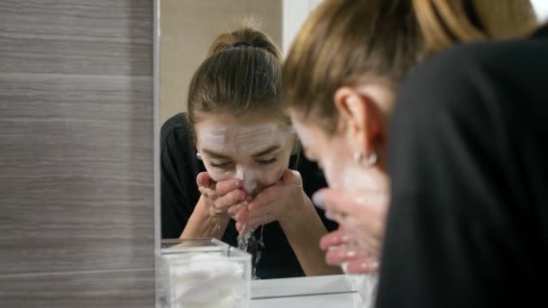 het meisje in het bad in de buurt van de spiegel, wassen van haar gezicht, wegspoelt het cosmetische masker. - Video