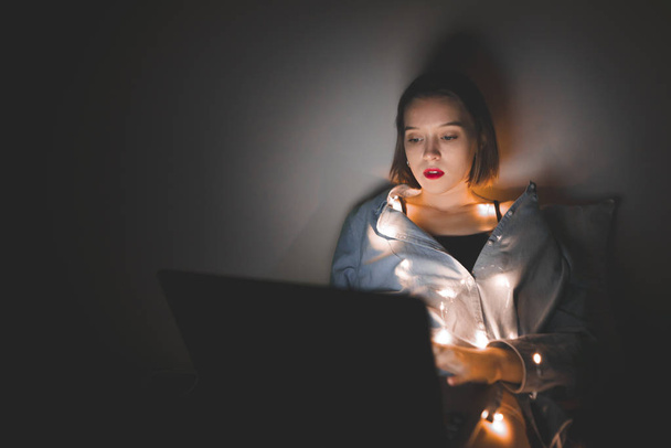 Woman with a laptop on her legs, lying in bed at Christmas lights at night, uses a laptop. Focus is on the girl hands works on a laptop at night. Gadget and technology concept. Night work - Zdjęcie, obraz
