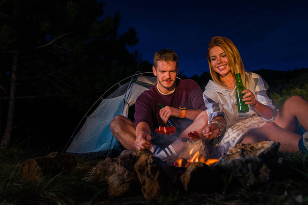 Paar bakken en eten van worst, bier drinken en ontspannen door het kampvuur in het bos in de nacht. Romantische kampvuur concept. - Foto, afbeelding
