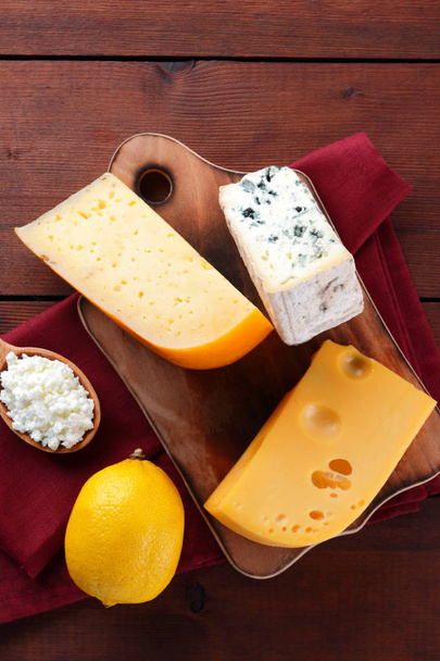 Queso duro, queso azul y requesón sobre una tabla de madera. Diferentes tipos de queso en una servilleta roja. Productos lácteos y limón sobre fondo de madera. Vista superior
 - Foto, imagen
