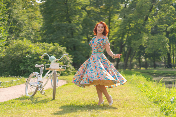 cute Caucasian girl with red hair in a retro dress enjoying life with a white vintage Bicycle in sporting fashionable walk in a city Park at summer day - Foto, immagini
