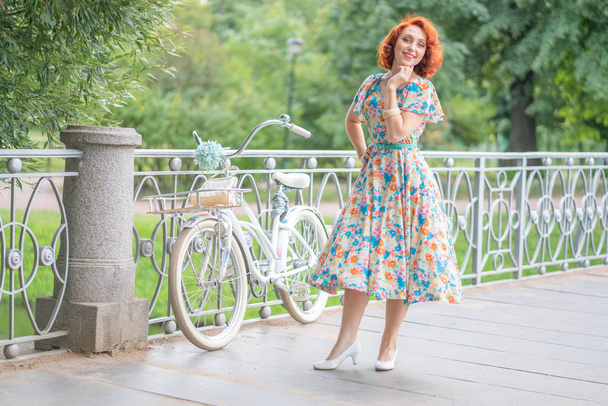 carino caucasico ragazza con i capelli rossi in un abito retrò godersi la vita con un bianco vintage Bicicletta in sport alla moda a piedi in un parco cittadino al giorno d'estate
 - Foto, immagini