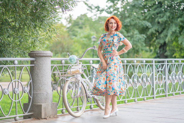 cute Caucasian girl with red hair in a retro dress enjoying life with a white vintage Bicycle in sporting fashionable walk in a city Park at summer day - Photo, Image