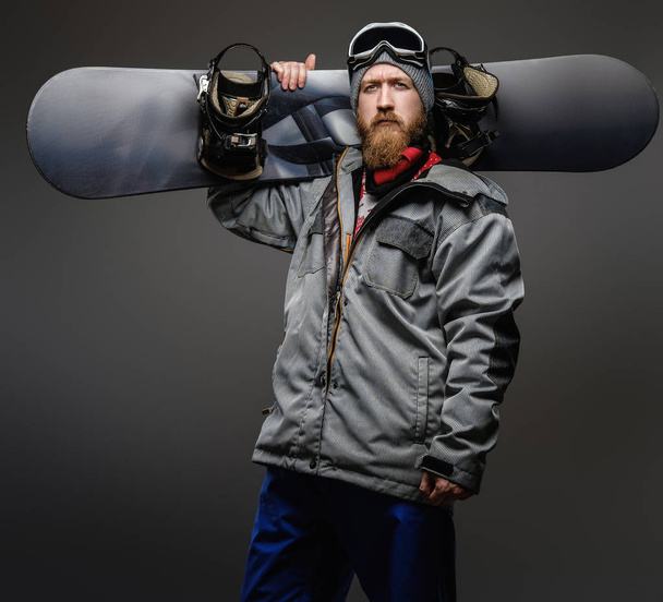 Confident man with red beard wearing a full equipment holding a snowboard on his shoulder, isolated on a dark background. - Fotoğraf, Görsel