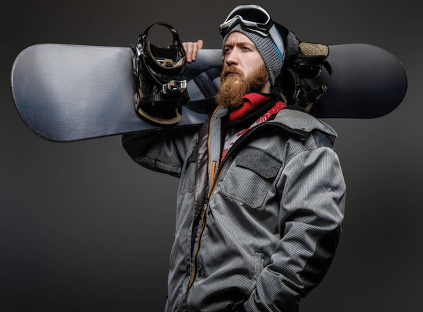 Zelfverzekerde man met een rode baard dragen van een volledige uitrusting met een snowboard op zijn schouder, op zoek weg met een serieuze blik, geïsoleerd op een donkere achtergrond. - Foto, afbeelding