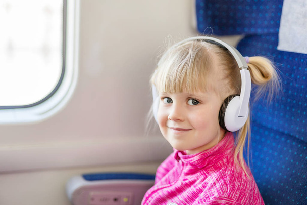 Little cute girl travelling by train. Kid listening to music with white headphones. Children activity and entertainment during trip concept - Fotó, kép