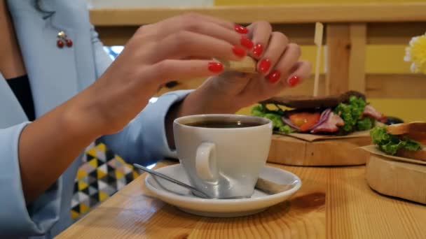 close-up of girl's hand with red manicure breaks sugar stick in the middle and pours sugar into a cup with fresh coffee standing on a wooden table next to sandwiches - Filmati, video