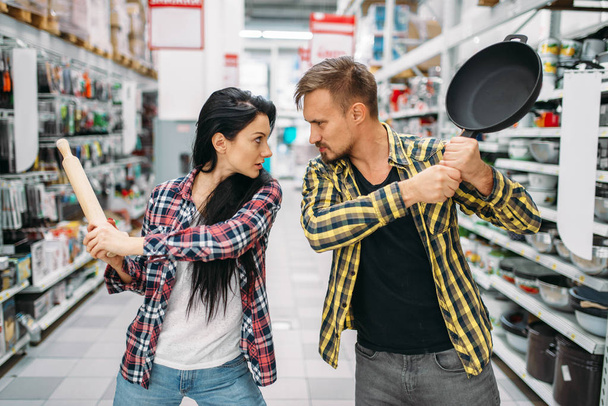 Young couple plays with frying pan and rolling pin in supermarket. Male and female customers on family shopping. Man and woman purchasing goods for the house - Photo, Image
