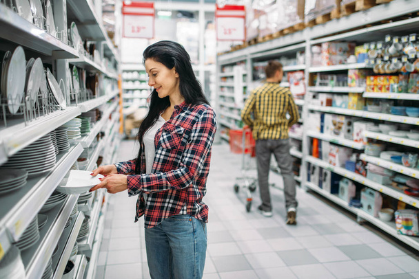 Молода жінка вибирає тарілки в супермаркеті. Жіночий клієнт на покупки в гіпермаркеті, продукти для кухні
 - Фото, зображення