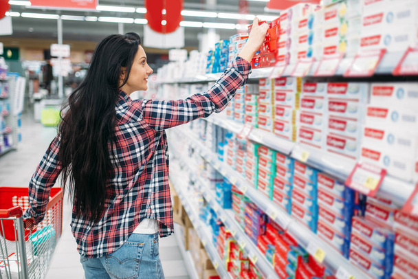 Молодая женщина покупает зубную пасту в супермаркете. Женщина-клиент по шопингу в гипермаркете, отдел товаров для личной гигиены
 - Фото, изображение