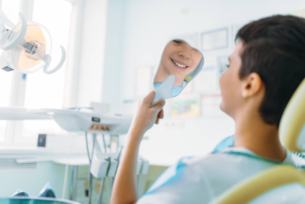 kleiner Junge im Zahnarztstuhl, der im Spiegel seine Zähne betrachtet, Kinderzahnheilkunde, Kinderstomatologie - Foto, Bild