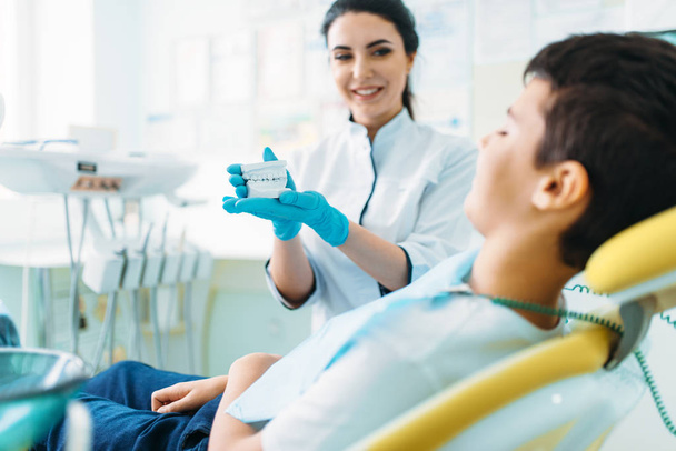 Γυναίκα οδοντίατρος δείχνει οδοντοστοιχίες να μικρό αγόρι σε οδοντιατρική καρέκλα, επαγγελματικής Παιδοδοντιατρικής, παιδιά Στοματολογία - Φωτογραφία, εικόνα