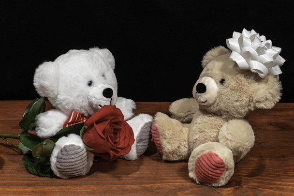 Twee schattige pluchen beren van teddy met enkele rode roos Ien witte strik op houten tafel op donkere achtergrond. Valentines, moeders dag, Pasen, Kerstmis, bruiloft concepten - Foto, afbeelding