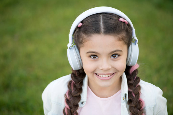 Listen music while relaxing outdoor. Kid girl enjoy music while sit on green grass meadow. Pleasant leisure time. Child headphones listen music. Fashion pretty cool girl in headphones listening music - Foto, afbeelding