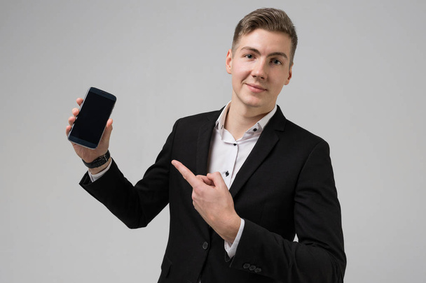 Nuoren miehen muotokuva mustassa puvussa, joka näyttää sormea matkapuhelimessa, jossa tyhjä näyttö on eristetty valkoisella taustalla
 - Valokuva, kuva