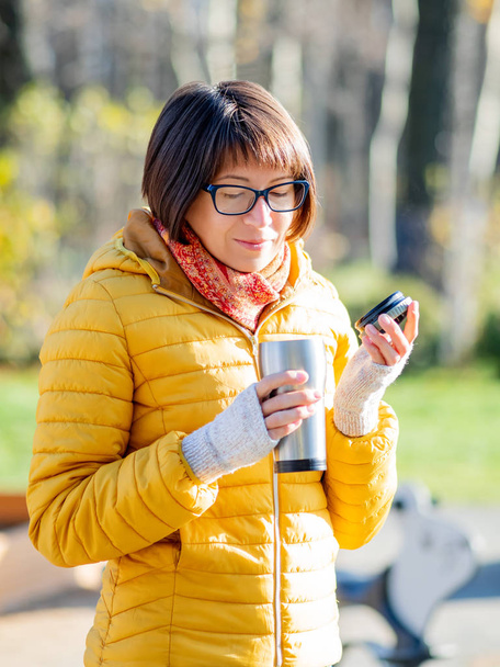 Happy wide smiling women in bright yellow jacket is holding thermos mug. Hot tea or other beverage on cool autumn day. - Photo, Image