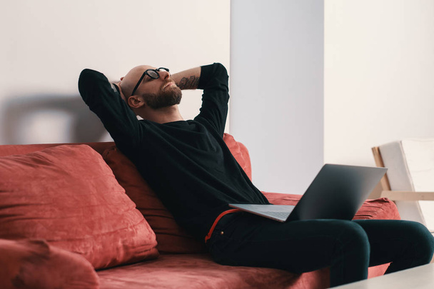moderner Mann sitzt auf Couch mit Laptop und entspannt sich nach anstrengender Arbeit mit den Händen hinter dem Kopf - Foto, Bild