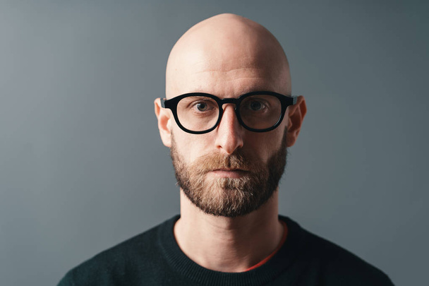 Крупним планом портрет впевнений стильний молодий бородатий чоловік в окулярах з чорною рамкою, позує на сірому фоні студії
. - Фото, зображення