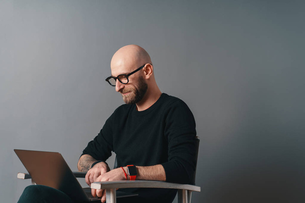 Сучасний білий чоловік з бородою і окулярами розслабляється в інтер'єрі студії, сидячи на срібному стільці, сірому фоні і серфінгу в Інтернеті на ноутбуці. Хлопець насолоджується онлайн-спілкуванням за допомогою гаджета
. - Фото, зображення