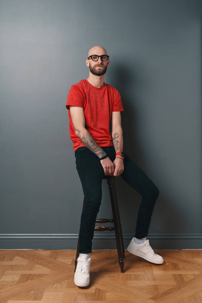 Vertrouwen stijlvolle jonge bebaarde man met bril met zwart frame, poseren in studio o grijze achtergrond zittend op een hoge stoel. - Foto, afbeelding