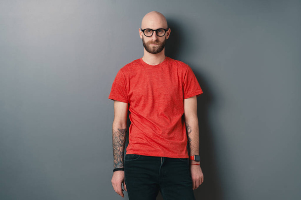 Впевнений стильний молодий бородатий чоловік в окулярах з чорною рамкою, позує на сірому фоні студії
. - Фото, зображення