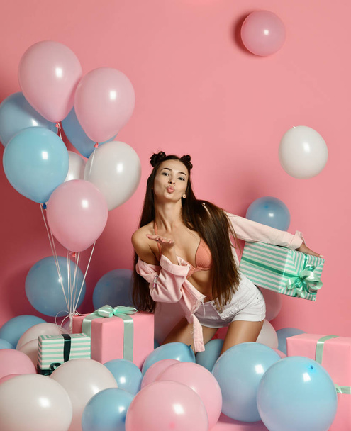Portrait of young woman in birthday party holding gift box with gift present on pastel pink background with colorful air baloons. - Fotó, kép