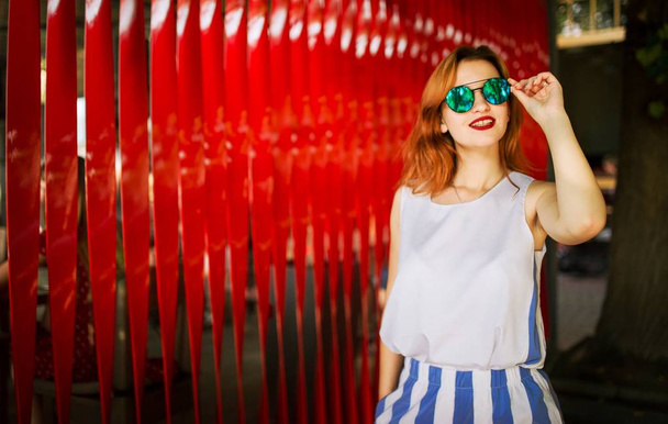 Attractive redhaired woman in sunglasses posing against red background. - Foto, imagen