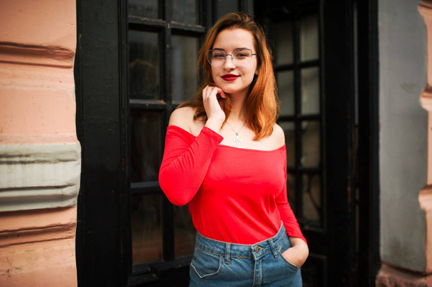 Attractive redhaired woman in eyeglasses, wear on red blouse and jeans skirt posing at street against old wooden door. - Foto, Imagen
