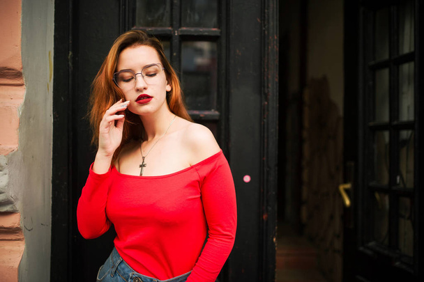 Attractive redhaired woman in eyeglasses, wear on red blouse and jeans skirt posing at street against old wooden door. - Zdjęcie, obraz